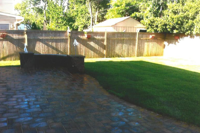 Patio with brick pavers, green lawn, wooden fence, trees, and a building in the background.