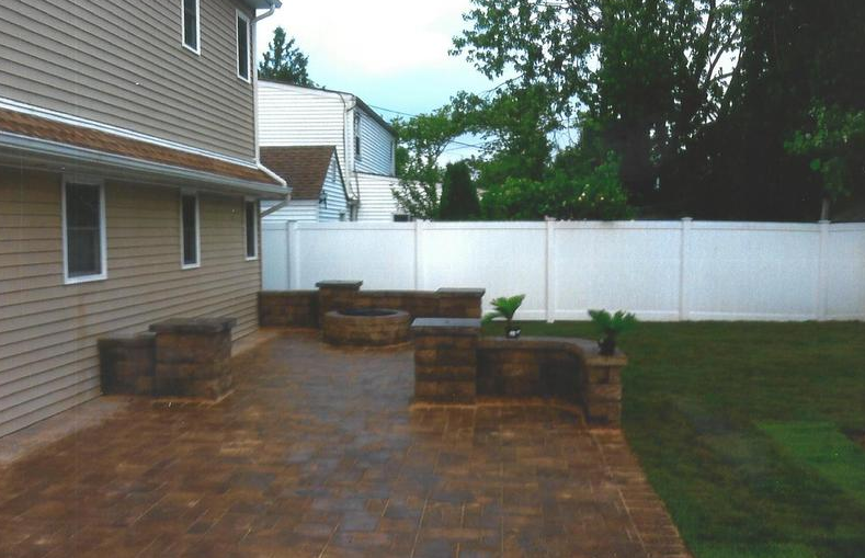 Brick patio with stacked stone features, fire pit, and beige house with a white fence in the background.