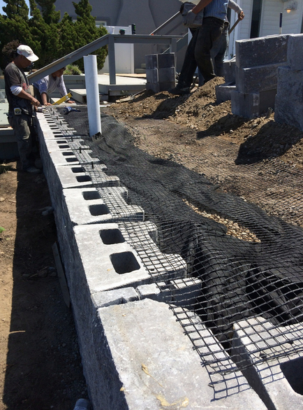 Construction workers building a retaining wall with concrete blocks, black mesh, and soil outdoors.