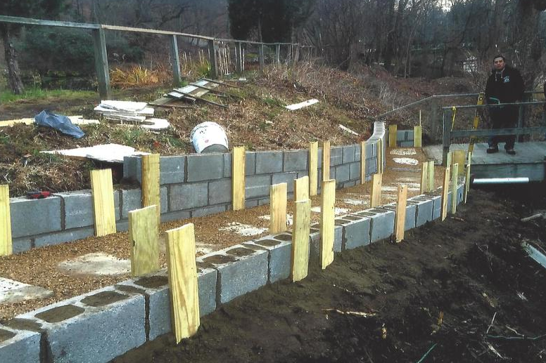A man builds a retaining wall with cinder blocks. Wooden supports hold blocks in place. Outdoor setting.