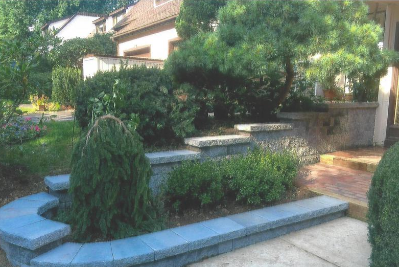 Stone retaining walls with tiered levels, lush greenery in front of a house.