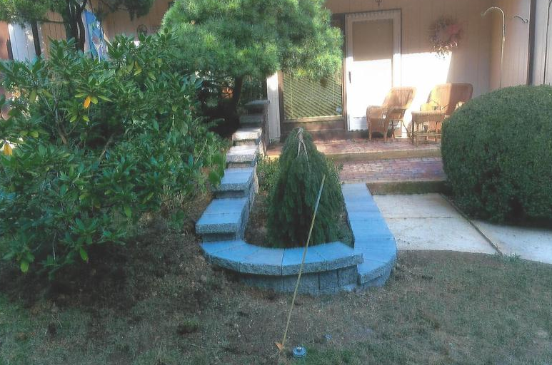 Stone-bordered garden bed with a small evergreen tree, located in front of a building and porch, on a grassy lawn.