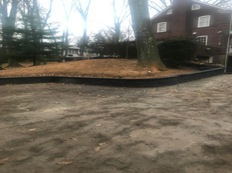 A brown building and a large tree next to a gravel driveway. There's a raised garden bed with brown foliage.