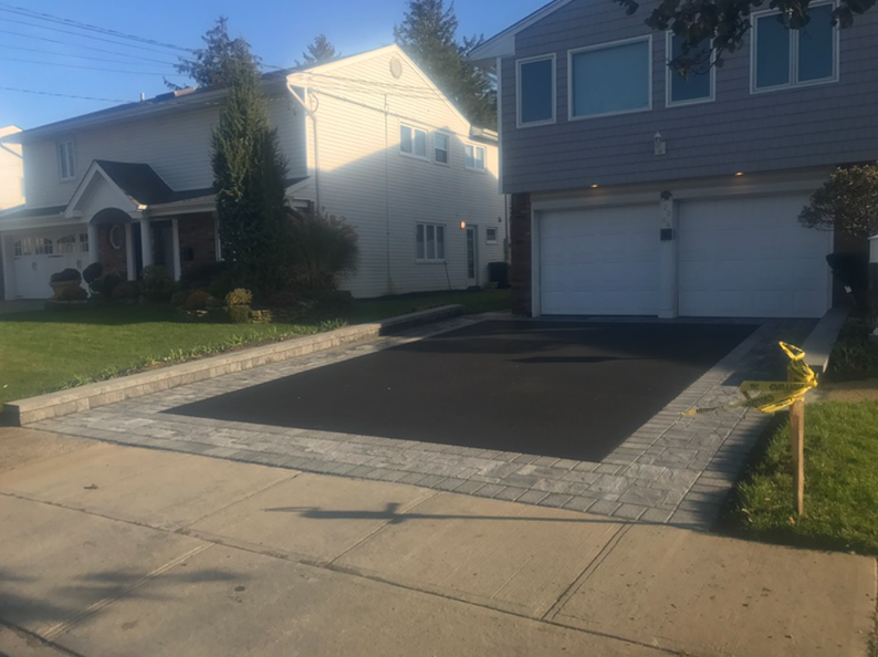 Driveway with blacktop bordered by gray pavers, leading to a two-car garage. Houses in the background.