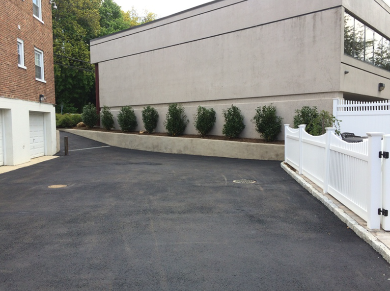 Asphalt parking area with low shrubs and white fence. Tan building on right, brick building on left.