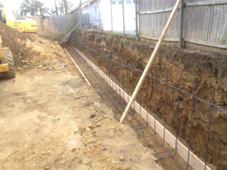 Trench with wooden forms and rebar, ready for concrete, next to a fence, in a yard.