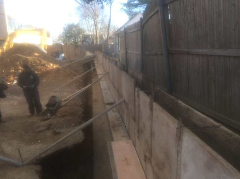 Construction site with concrete retaining wall, fence, and workers. Earth and equipment in background.