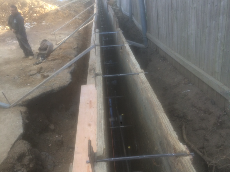 Construction of a narrow concrete foundation trench with rebar, next to a wooden fence and workers.