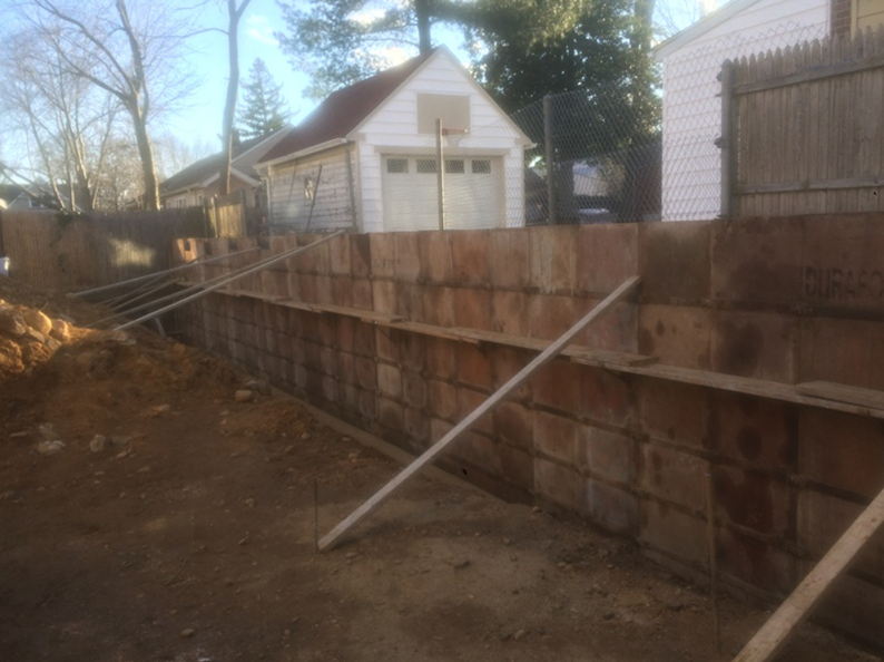 Construction site with a brown retaining wall, dirt ground, and a white garage in the background.