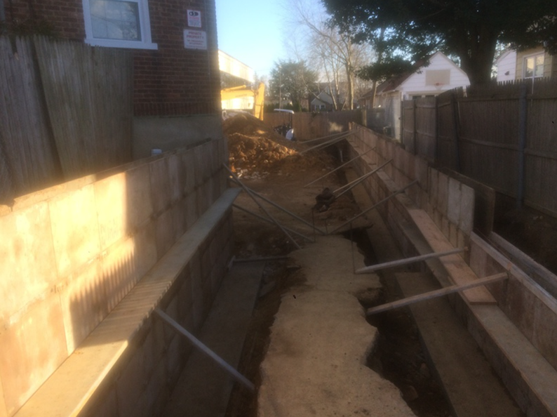 Construction site with trenches, wooden supports, and an excavator in the background.
