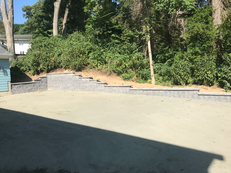 A paved area with retaining walls and a slope, surrounded by trees; the sky is clear.