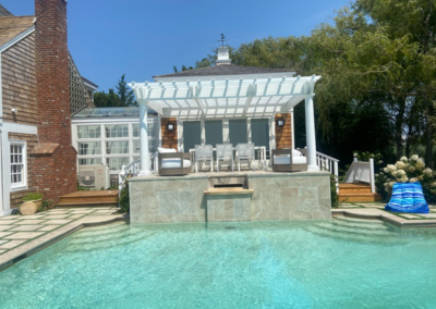 Poolside pergola with table and chairs; building with brick and wood siding in background.