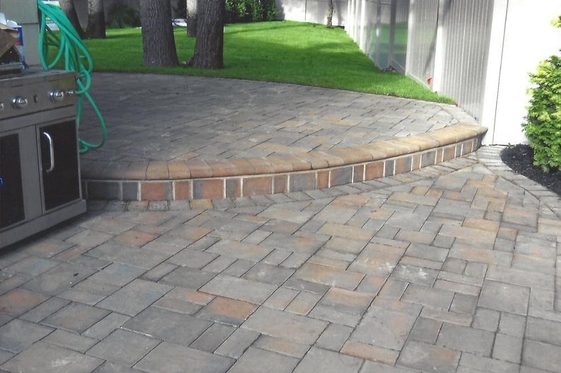 Curved brick patio with a raised border, adjacent to grass and trees, next to an outdoor kitchen and a fence.