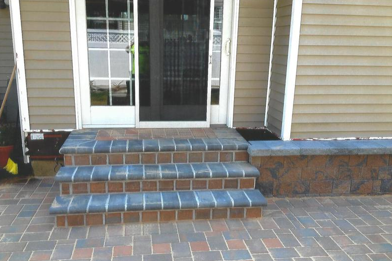 Three brick steps lead to a glass door of a house. The steps and patio are brick, and the house is beige.