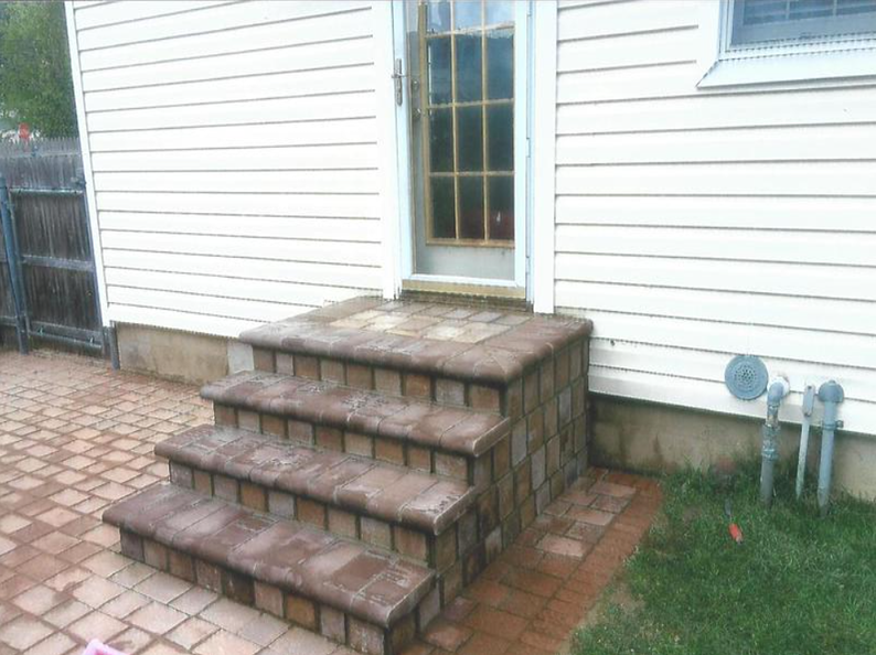 Brick steps leading up to a glass door on a white-sided house, surrounded by brick and grass.