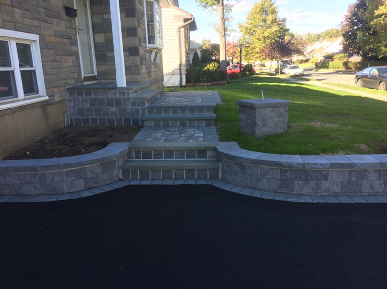 A paved stone walkway with steps leads to a house's front door, surrounded by a low retaining wall and lawn.