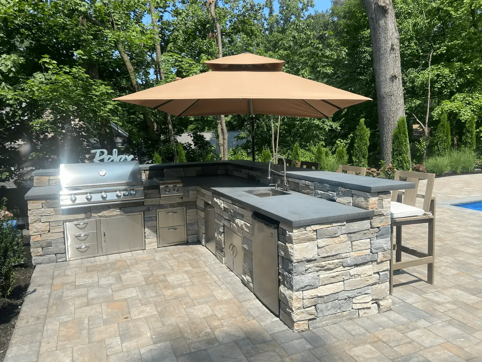 Outdoor kitchen with stone facade, grill, sink, and seating under a tan umbrella.