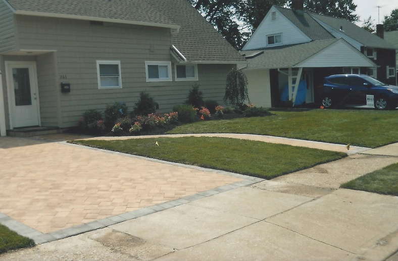 House with beige driveway, green lawn, and flower bed. Another house and car are visible.