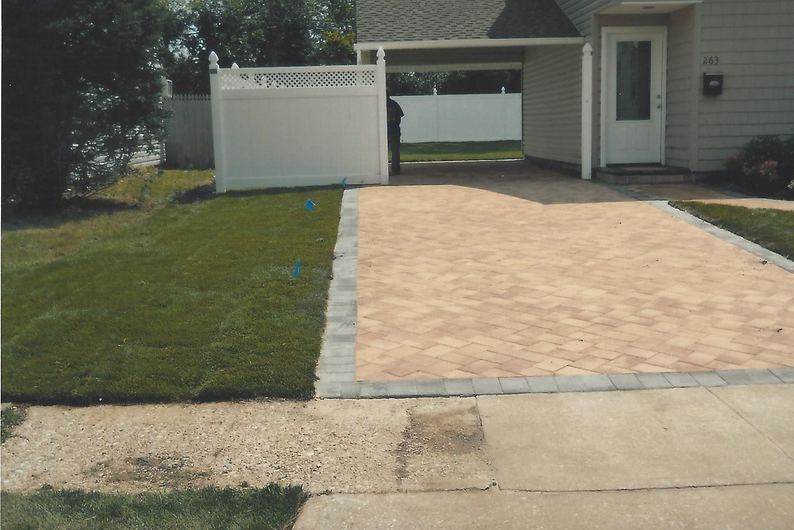 Driveway paved with tan bricks, bordered by gray bricks, beside green lawn and white fence.