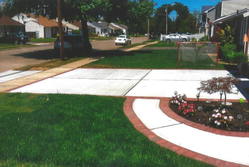 A residential street with a green lawn, white concrete driveway, and a flowerbed. A car drives on the street.
