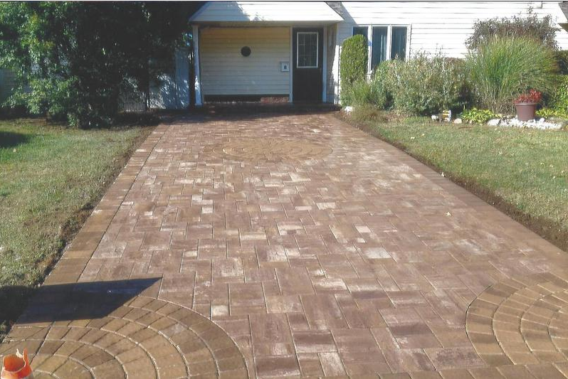 Brick driveway leading to a house with a garage. Brown bricks, green lawn.