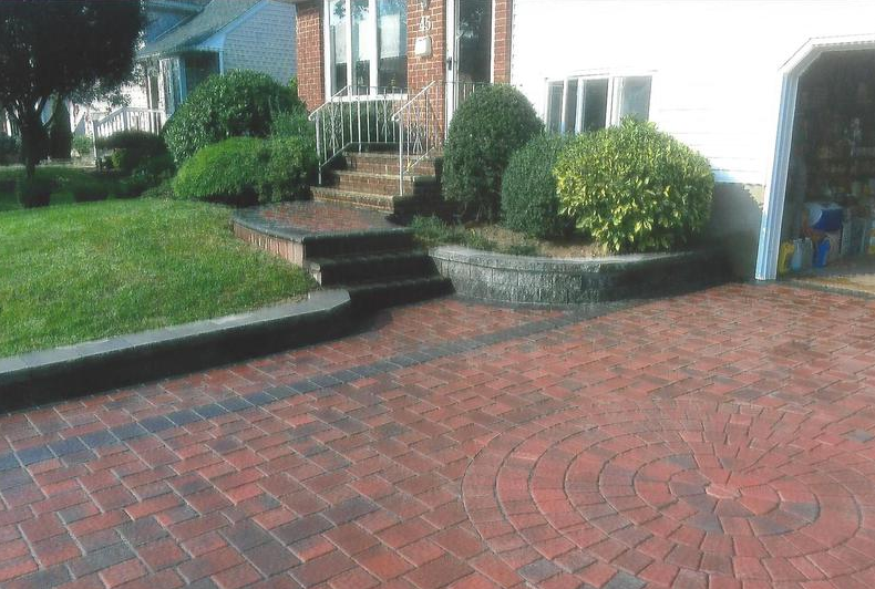 Brick driveway with circular pattern, low brick wall, green bushes, and lawn in front of a house.