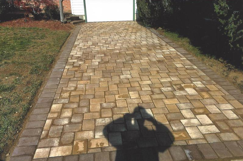 Driveway paved with brown and tan bricks; shadow of person holding a camera.