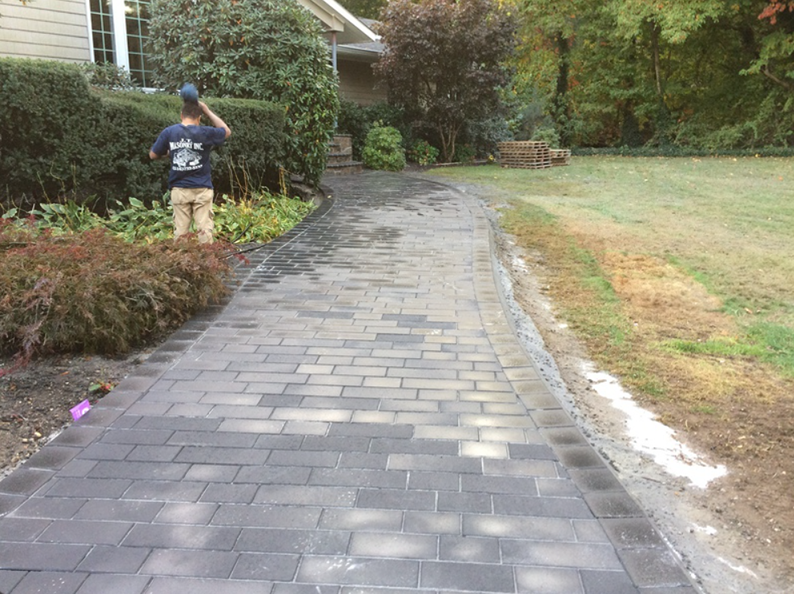 Brick walkway leading to a house, with a person in the background near bushes.