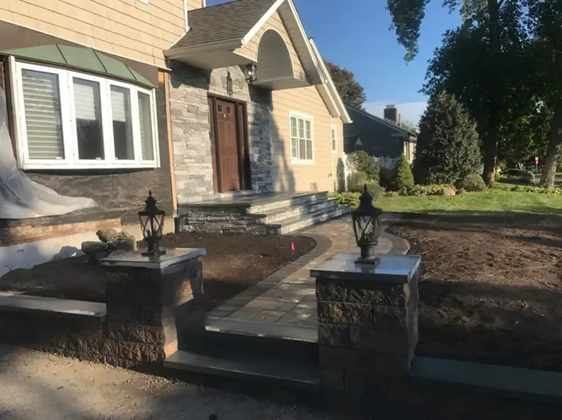 A house with stone steps and walkway, two lamp posts, and a grassy lawn on a sunny day.