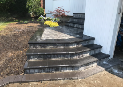 Stone steps leading to a house with a patio area, featuring landscaping with flowers and a small tree.