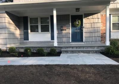 Front of house with blue door, stonework steps, gray siding, and shrubs.