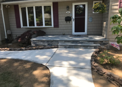 Concrete pathway leads to a house with stone steps.  House has beige siding and a white door.