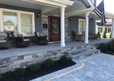 A house with a porch, stone steps, and dark blue siding. Landscaping with pavers and greenery. A flag is visible.