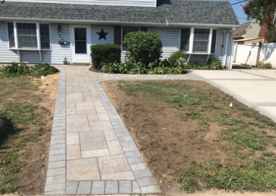 A brick pathway leads to a light blue house with a small yard, on a sunny day.