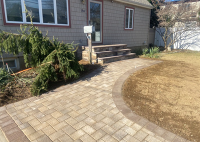 Brick pathway leads to a house entrance with steps. Beige bricks, small bushes, and a lawn.