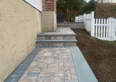 Brick steps and walkway with patterned pavers, leading up to a house next to a white fence.