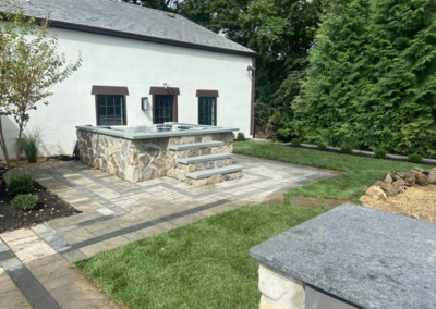 Stone-walled hot tub with steps on a paved patio next to a white building and grass.