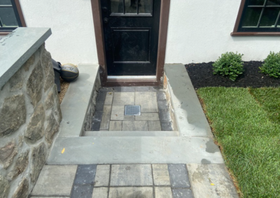 Black front door with steps. Stone and paving entrance. Dark mulch and green grass on the right.