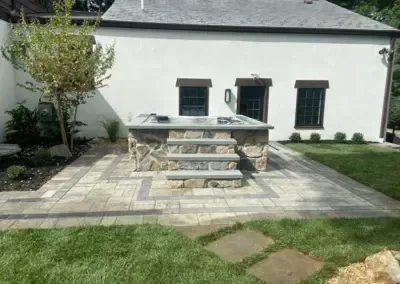 Stone hot tub with steps on a paved patio in front of a white building with dark trim.