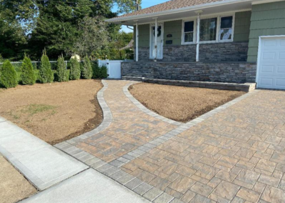 Brick walkway leads to a green house with stone siding; tan and brown pavers contrast with the green lawn.