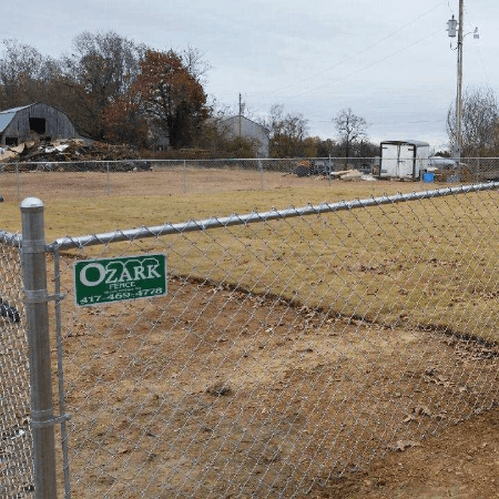 A chain-link fence with a company sign encloses a grassy field, rural setting with a barn and shed.