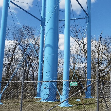 Blue water tower with metal support beams, fence, and trees against a blue sky.