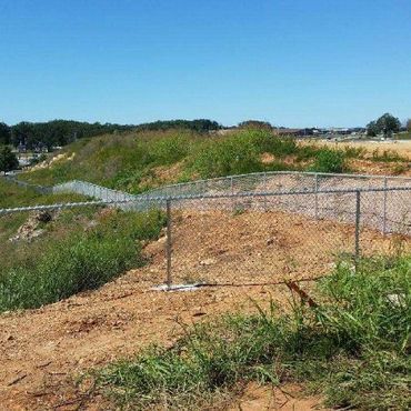 Chain-link fence bordering a dirt hill with green vegetation and a clear blue sky.