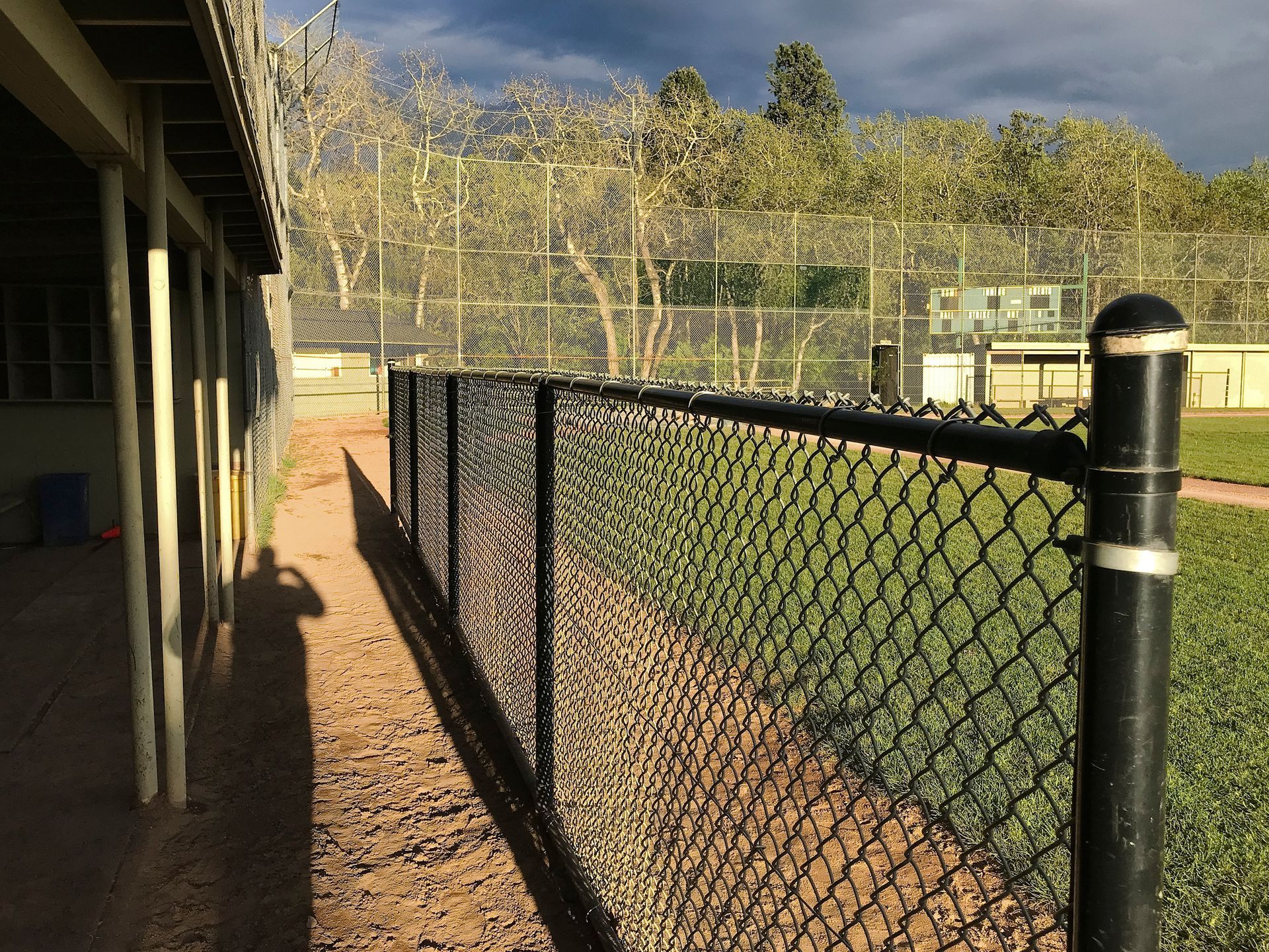 Baseball field with a black chain link fence, grassy area, and building with a shadow.