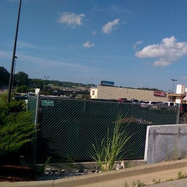 Green chain-link fence in front of a parking lot and buildings under a blue sky.