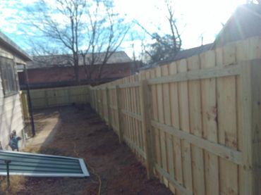 Wooden fence in a backyard, surrounding a dirt path. Trees and houses in the background, sunny day.