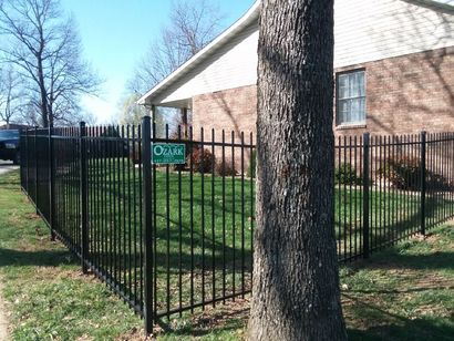 Black metal fence surrounding a green lawn next to a brick house and a tree.