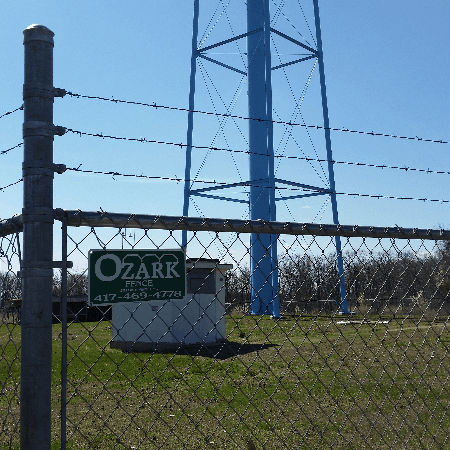 A blue water tower behind a chain-link fence with a sign for Ozark, Missouri.