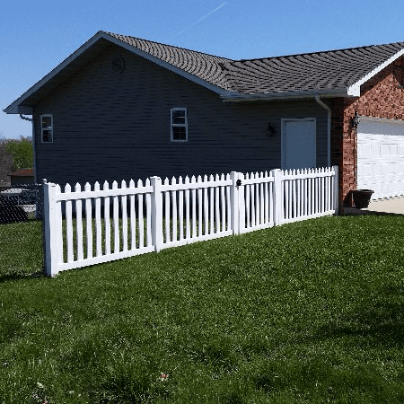 White picket fence in front of a gray and brick building, on green grass. Sunny day.