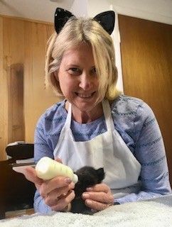 Woman in cat ears feeding a black kitten with a bottle, inside room.